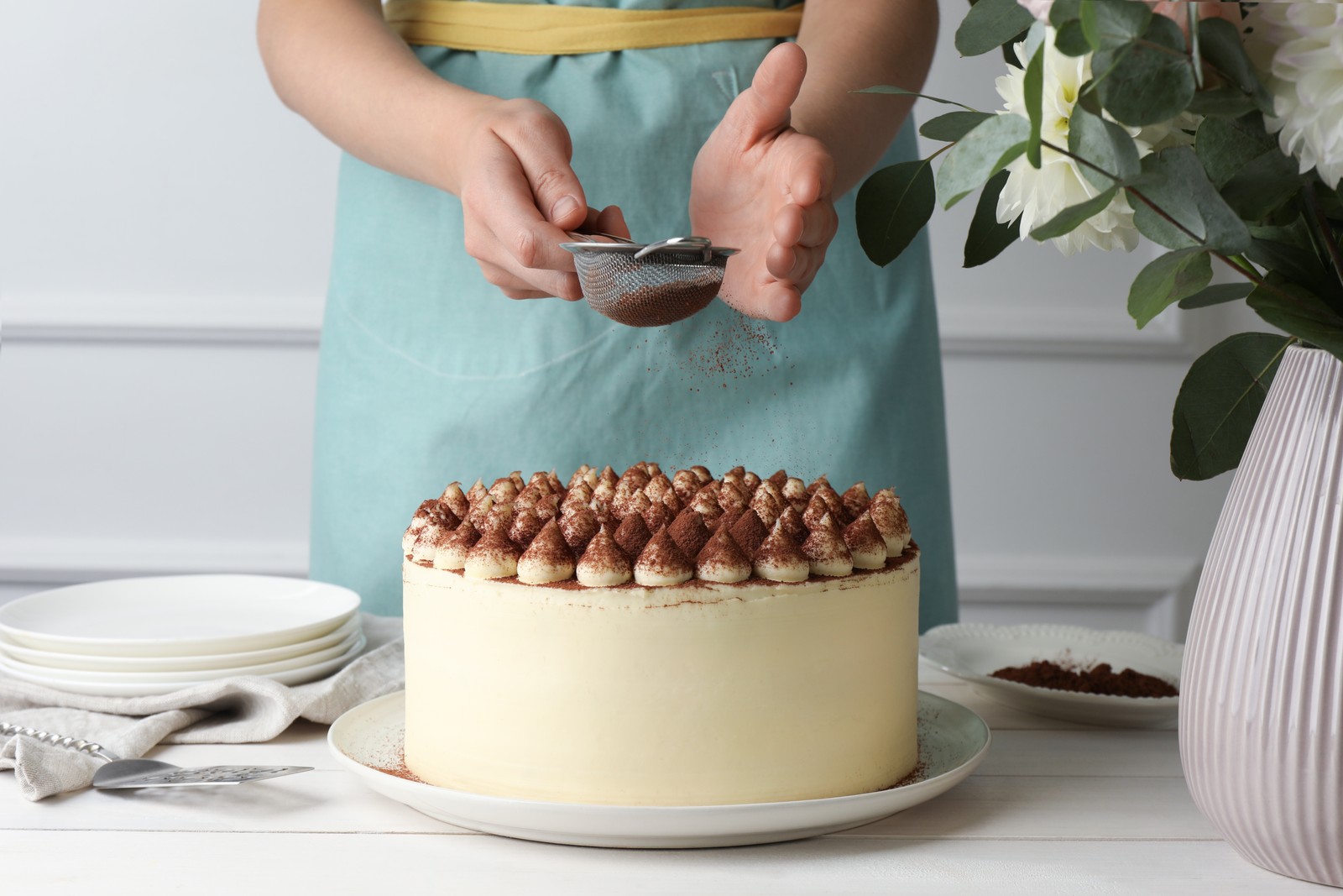 Photo of woman dusting delicious tiramisu cake with cocoa powder at white wooden table, closeup
