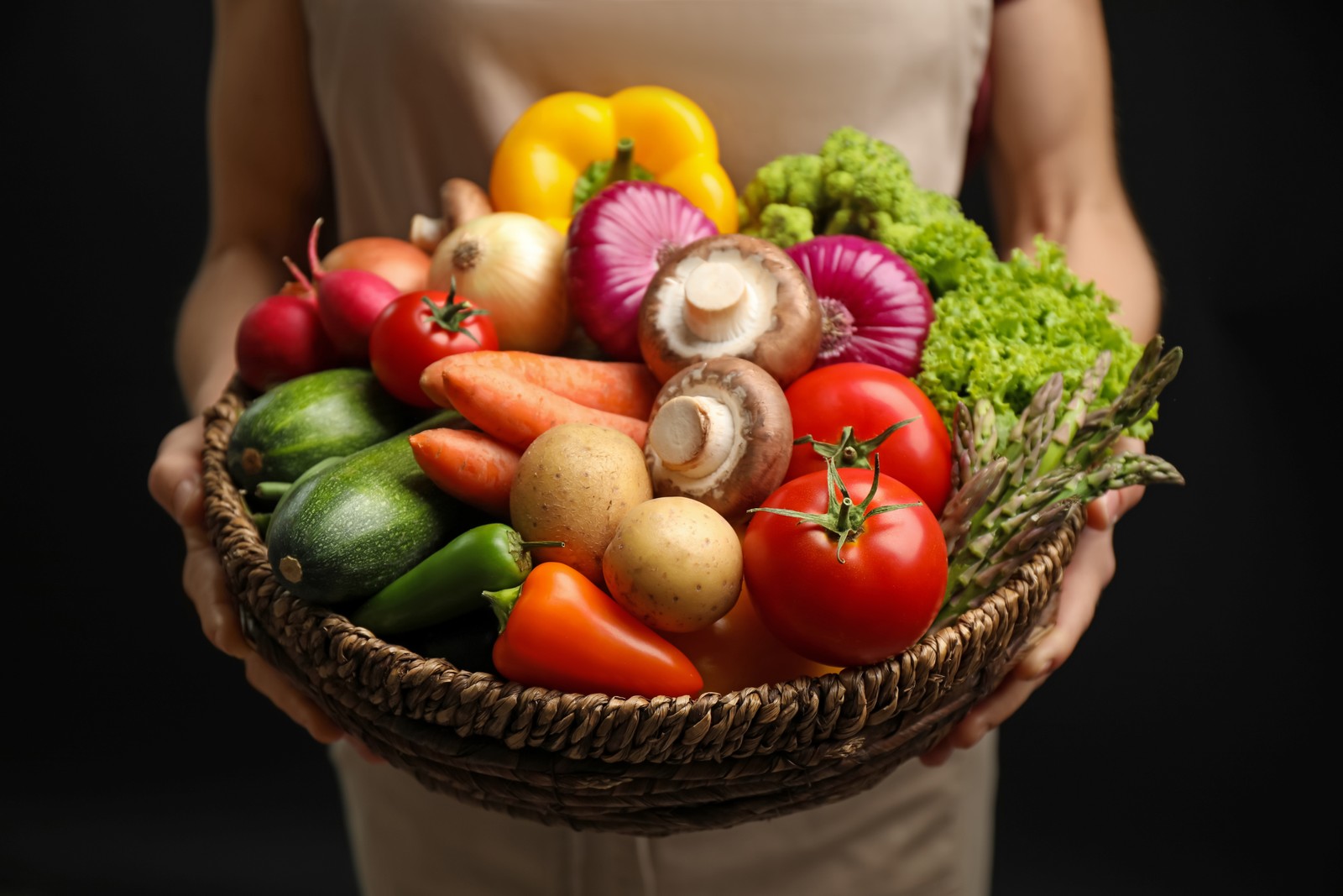 Photo of woman holding wicker basket full of fresh vegetables on black background, closeup