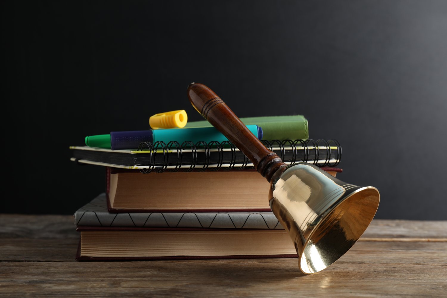 Photo of golden bell, books and stationery on wooden table near blackboard. School days