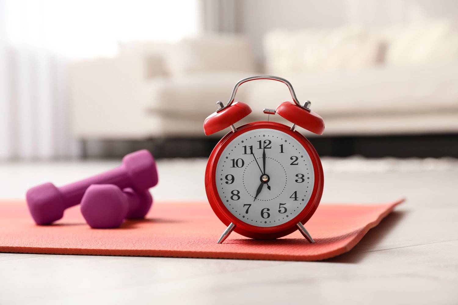 Photo of alarm clock, yoga mat and dumbbells on wooden floor indoors. Morning exercise