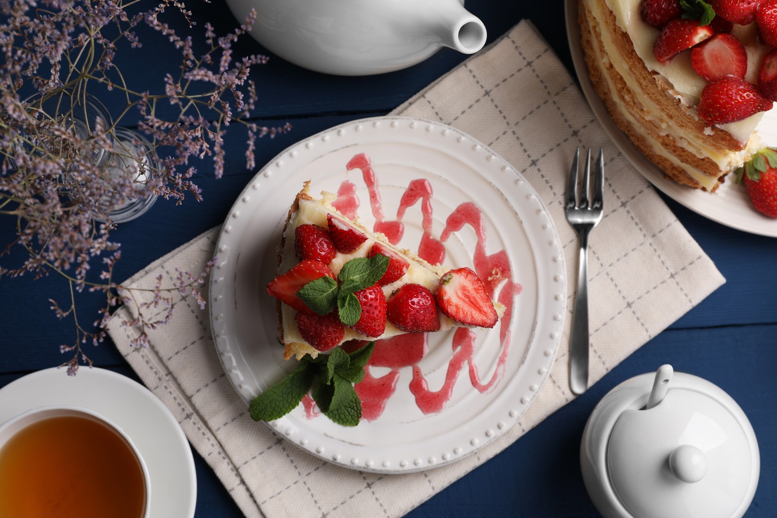 Photo of tasty cake with fresh strawberries, mint and cup of tea on blue wooden table, flat lay