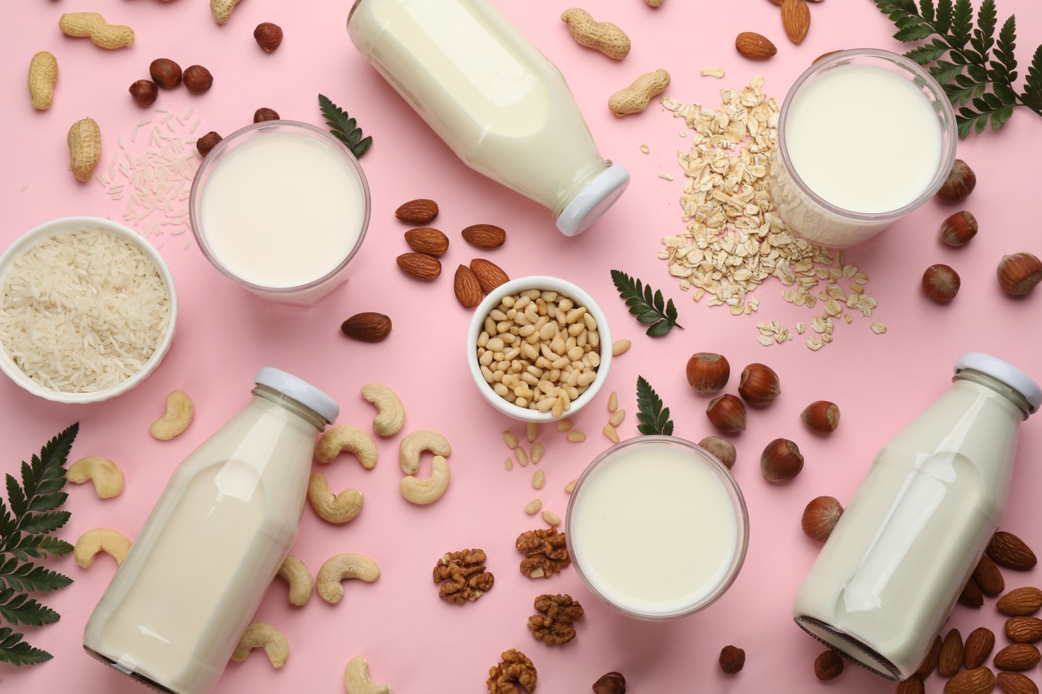 Photo of different vegan milks and ingredients on pink background, flat lay