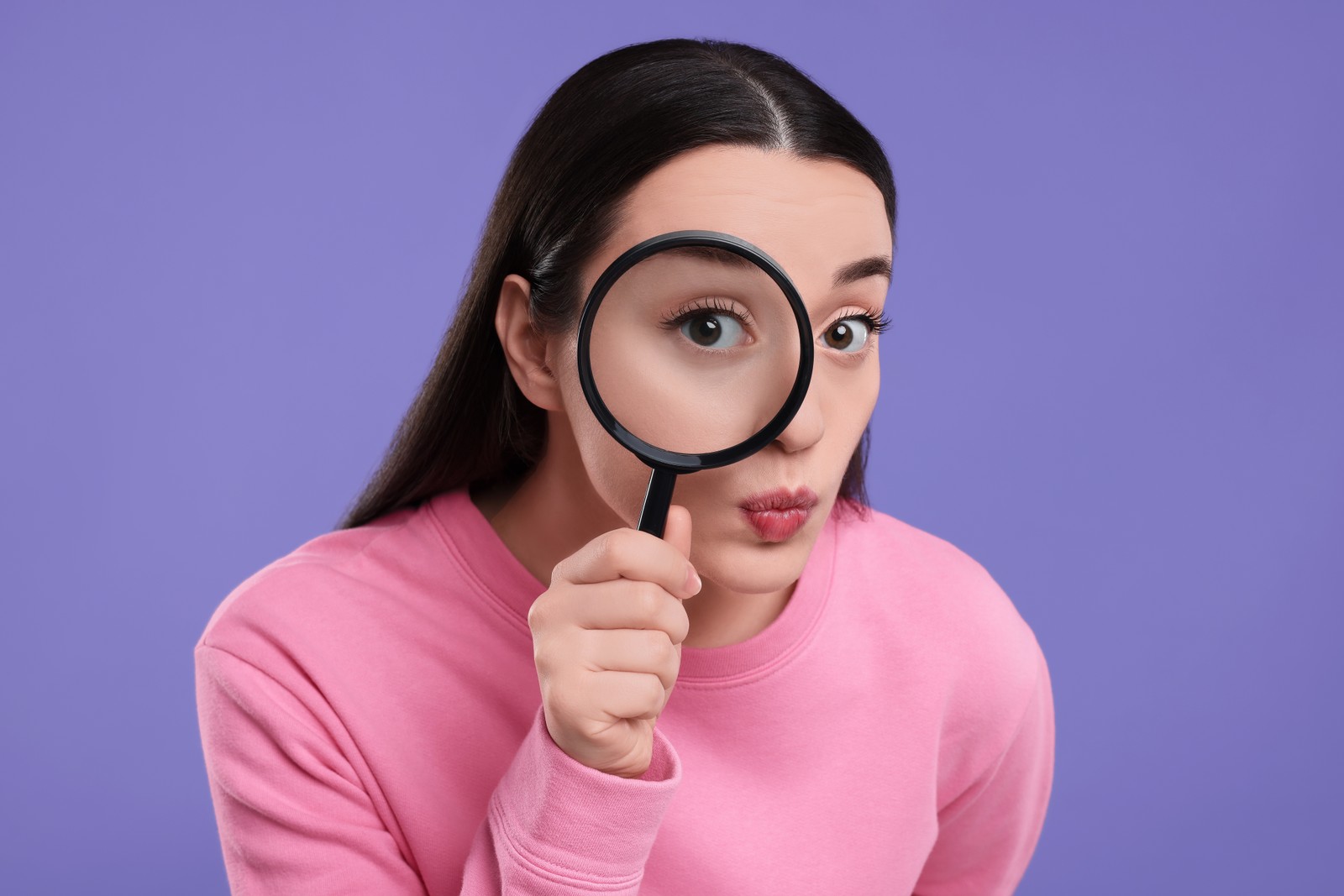 Photo of confused young woman looking through magnifier glass on purple background