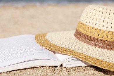 Photo of Open book and hat on sandy beach near sea, closeup