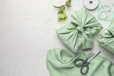 Photo of Furoshiki technique. Gifts packed in green fabrics, hellebore flowers, scissors and decorative ribbon on white wooden table, flat lay with space for text