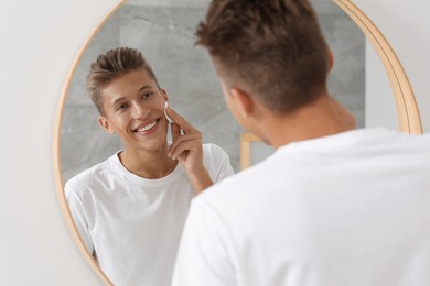 Photo of Handsome young man cleaning face with cotton pad near mirror in bathroom