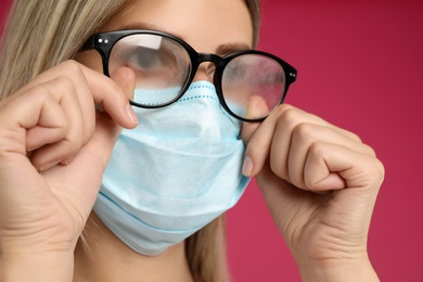 Photo of Woman wiping foggy glasses caused by wearing disposable mask on pink background, closeup. Protective measure during coronavirus pandemic