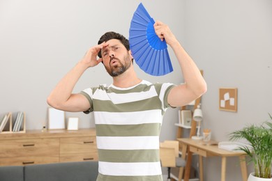 Photo of Bearded man waving blue hand fan to cool himself at home