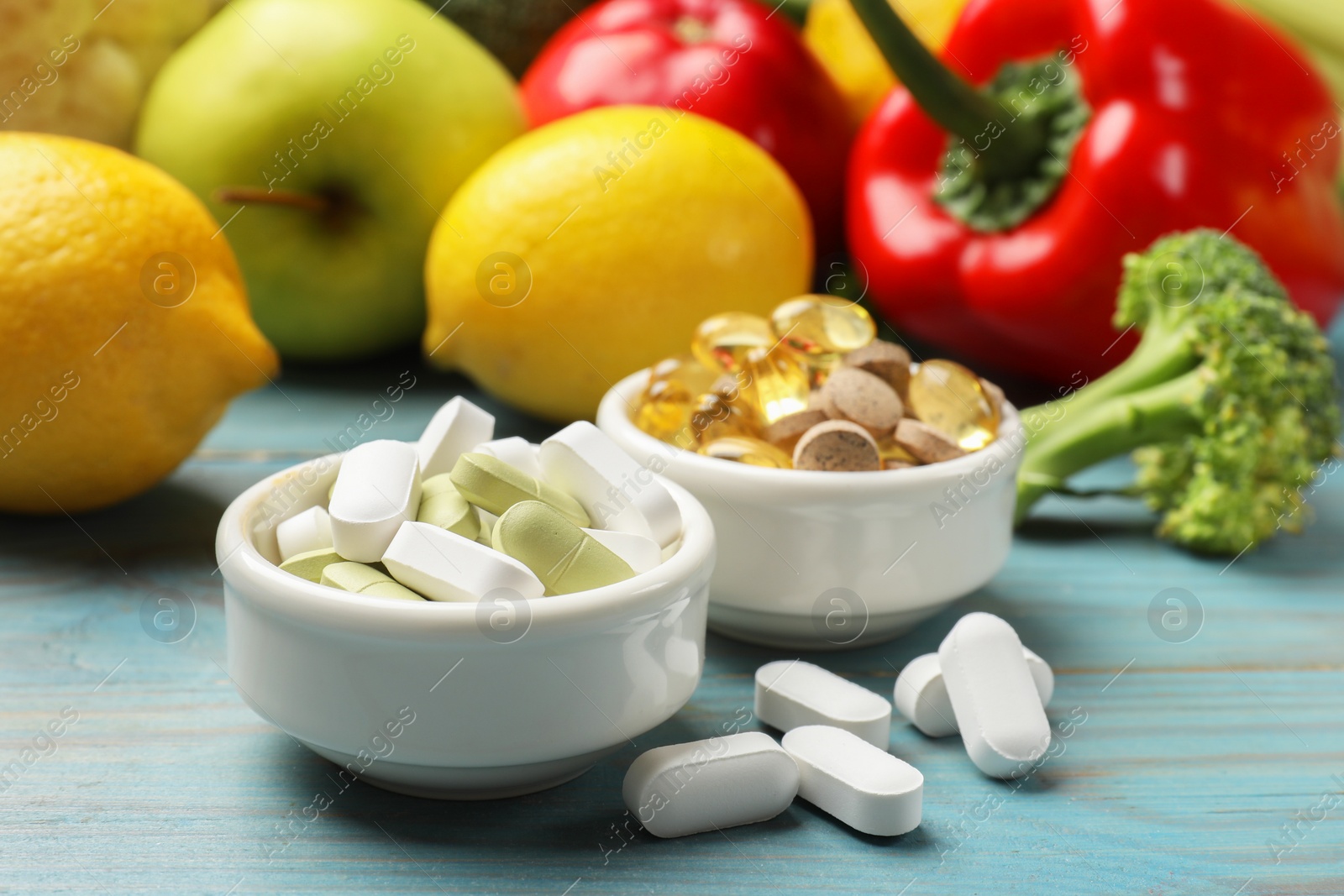 Photo of Dietary supplements. Bowls with different pills near food products on light blue wooden table, closeup