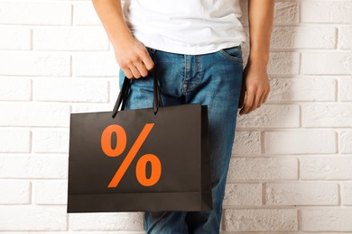 Image of Man holding paper shopping bag with percent sign near white brick wall, closeup. Discount concept