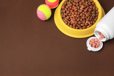 Photo of Bowl with dry pet food, bottle of vitamins and toys on brown background, above view. Space for text