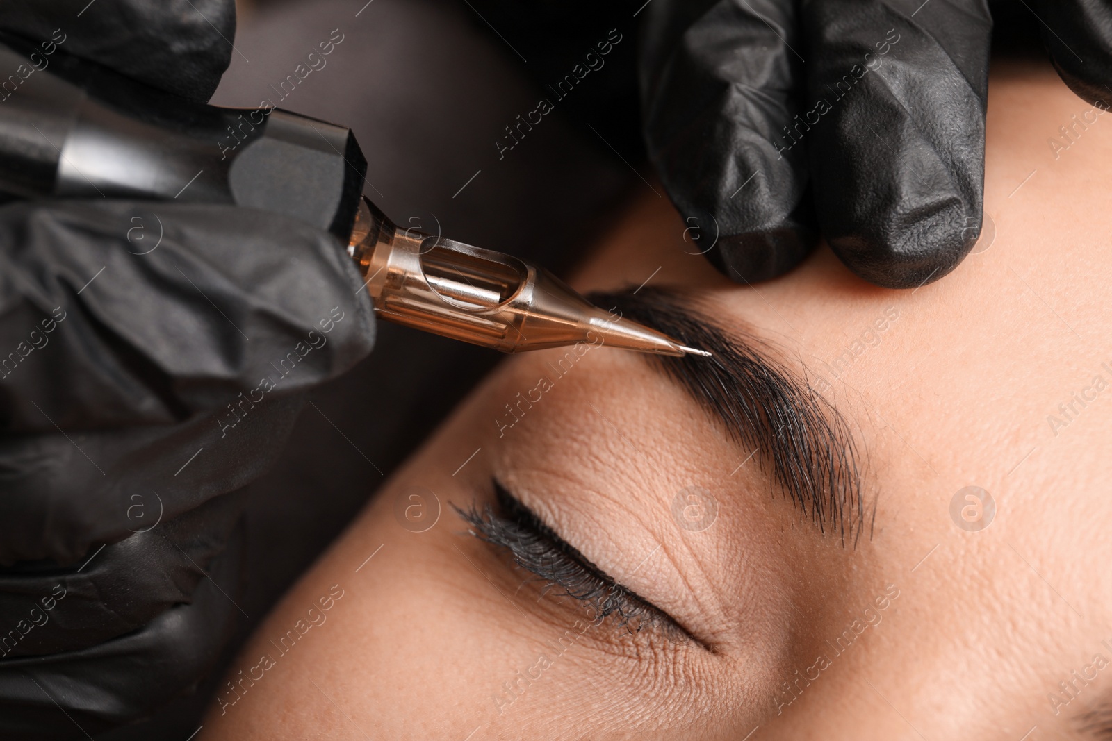 Photo of Young woman undergoing procedure of permanent eyebrow makeup in tattoo salon, closeup