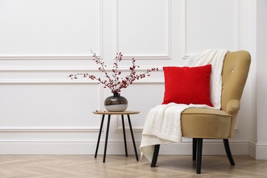 Photo of Hawthorn branches with red berries in vase on table near armchair indoors