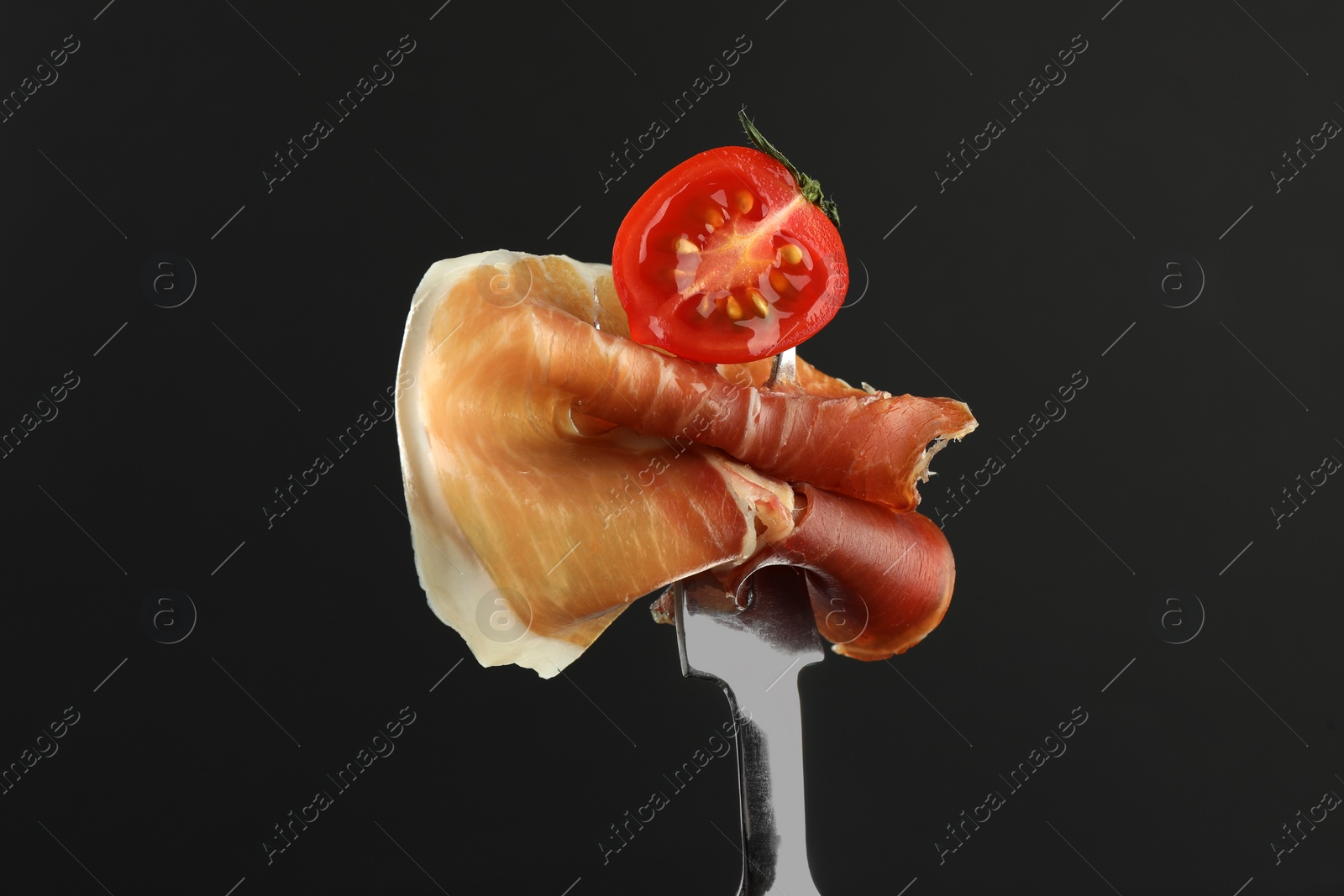 Photo of Fork with slice of tasty jamon and tomato on black background, closeup