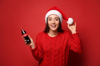 Photo of MYKOLAIV, UKRAINE - JANUARY 27, 2021: Young woman in Christmas hat holding bottle of Coca-Cola on red background