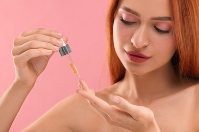 Photo of Beautiful young woman applying cosmetic serum onto her finger on pink background, closeup