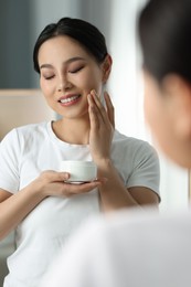 Photo of Happy woman applying face cream near mirror at home