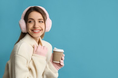 Happy woman with cup of drink wearing warm earmuffs on light blue background, space for text