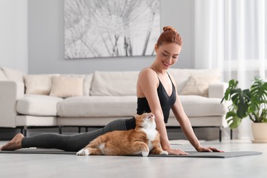 Beautiful woman with cute red cat practicing yoga on mat at home