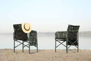 Camouflage fishing chairs with hat on sandy beach near river