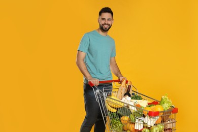 Photo of Happy man with shopping cart full of groceries on yellow background