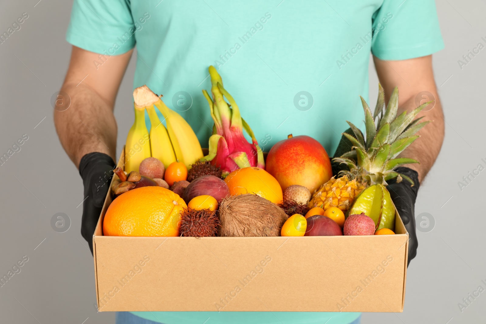 Photo of Courier holding box with assortment of exotic fruits on grey background, closeup