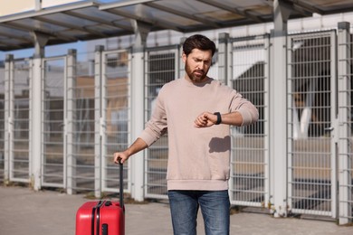 Photo of Being late. Worried man with red suitcase looking at his watch outdoors