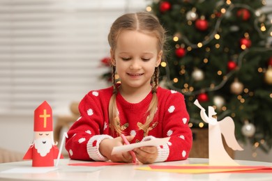 Photo of Cute little girl cutting paper at table with Saint Nicholas toy indoors