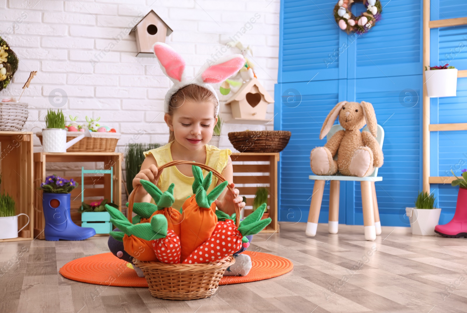 Photo of Adorable little girl with bunny ears and basket full of toy carrots in Easter photo zone