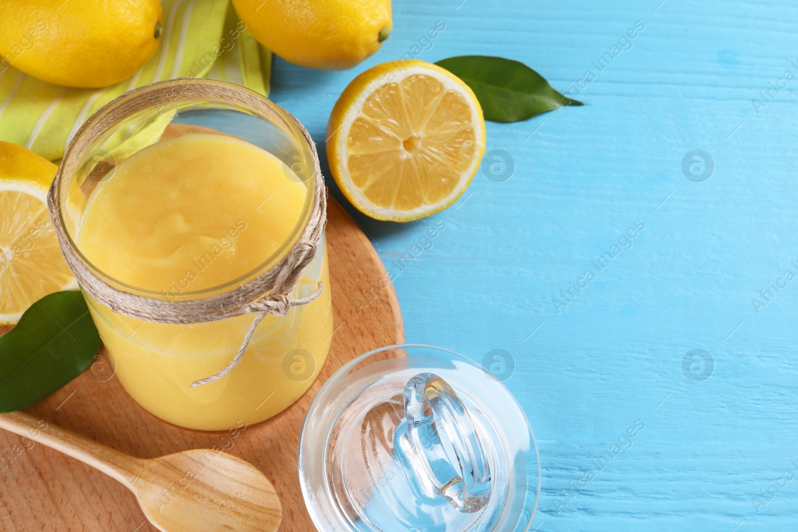 Photo of Delicious lemon curd in glass jar, fresh citrus fruits, spoon and green leaves on light blue wooden table, space for text