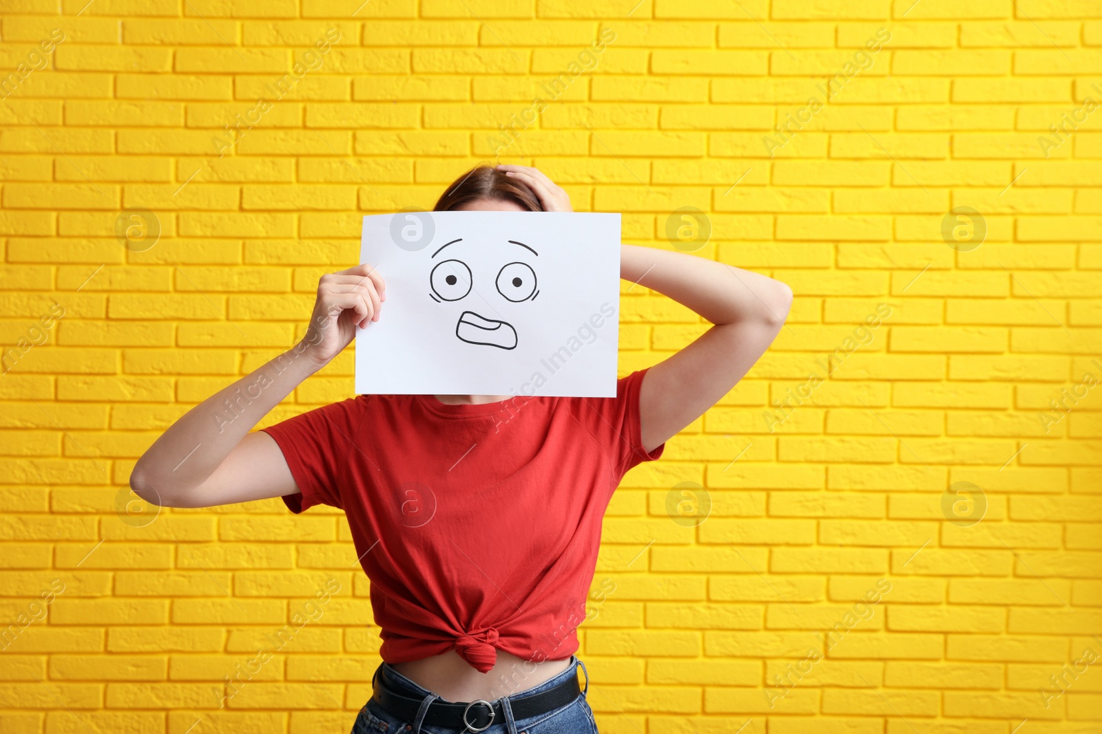 Photo of Woman hiding behind sheet of paper with scared face near yellow brick wall
