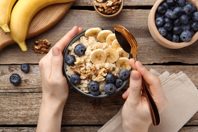 Photo of Woman eating tasty oatmeal with banana, blueberries and walnuts at wooden table, top view