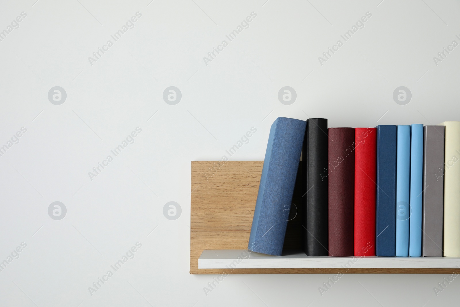 Photo of Many hardcover books on wooden shelf near white wall. Space for text