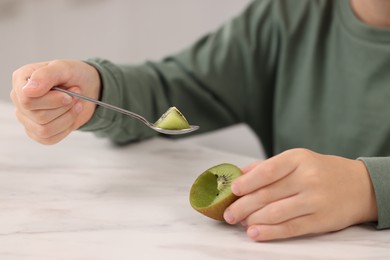 Boy eating tasty fresh kiwi with spoon at white marble table indoors, closeup