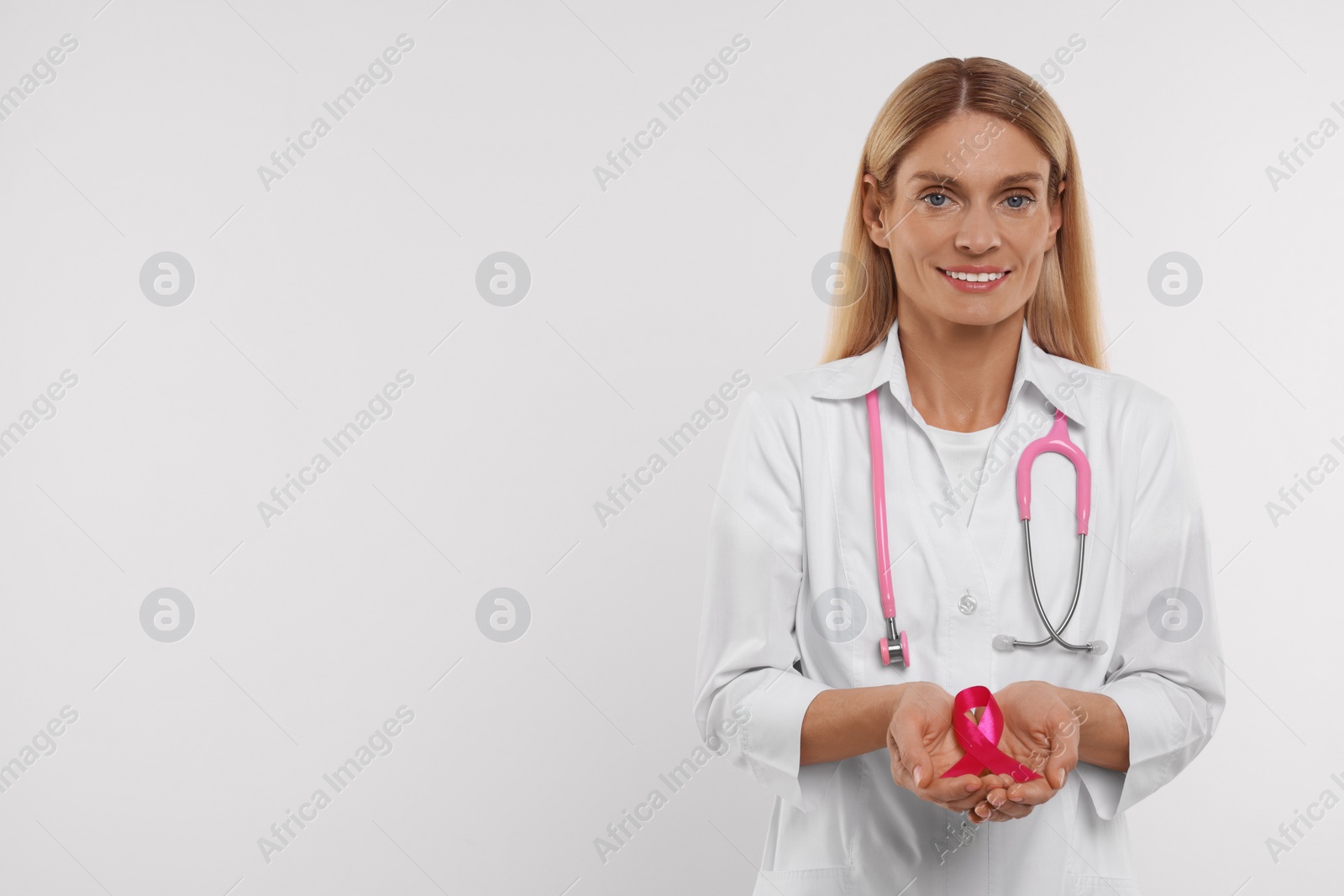 Photo of Doctor with stethoscope holding pink ribbon on white background, space for text. Breast cancer awareness