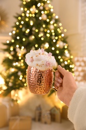 Photo of Woman holding cup of delicious drink with whipped cream and marshmallows near Christmas tree indoors, closeup