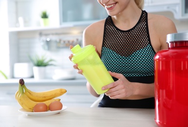 Young woman holding bottle of protein shake at table with ingredients in kitchen, closeup. Space for text