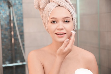 Photo of Beautiful woman with towel on head applying face cream in bathroom
