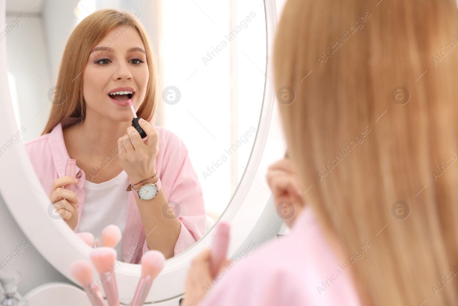 Photo of Beautiful young woman applying makeup near mirror indoors