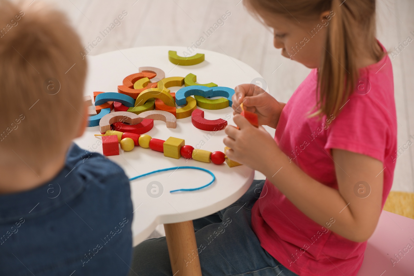 Photo of Little children playing with wooden pieces and string for threading activity at white table indoors. Developmental toys