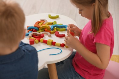 Little children playing with wooden pieces and string for threading activity at white table indoors. Developmental toys