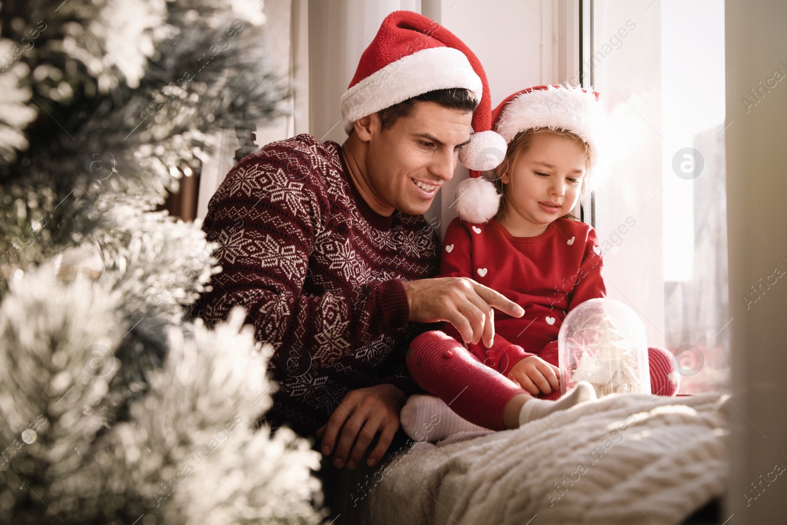 Photo of Father and daughter in Santa hats playing with snow globe near window