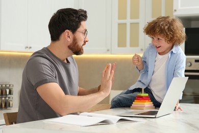Photo of Little boy bothering father at home. Child playing with toys on desk while his father working remotely