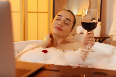 Woman with glass of wine taking bath in tub with foam and rose petals indoors
