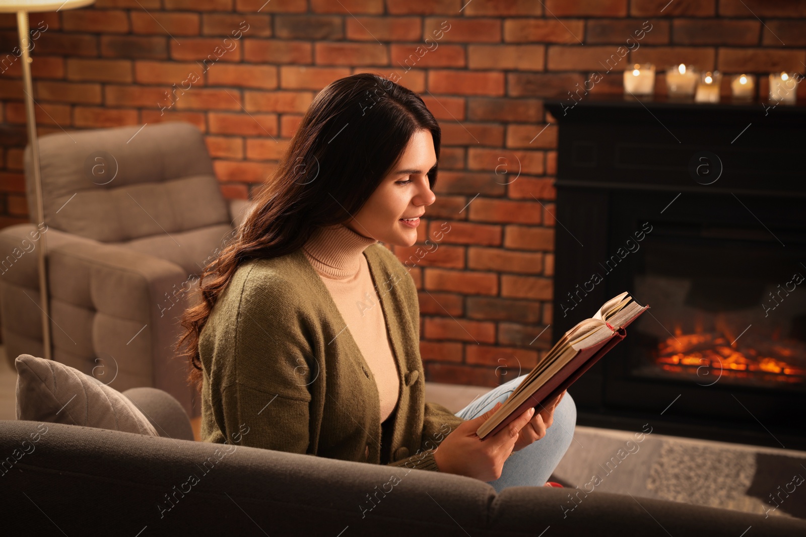 Photo of Young woman reading book on sofa near fireplace at home