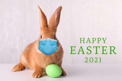 Image of Text Happy Easter 2021 and cute bunny in protective mask on light background. Holiday during Covid-19 pandemic