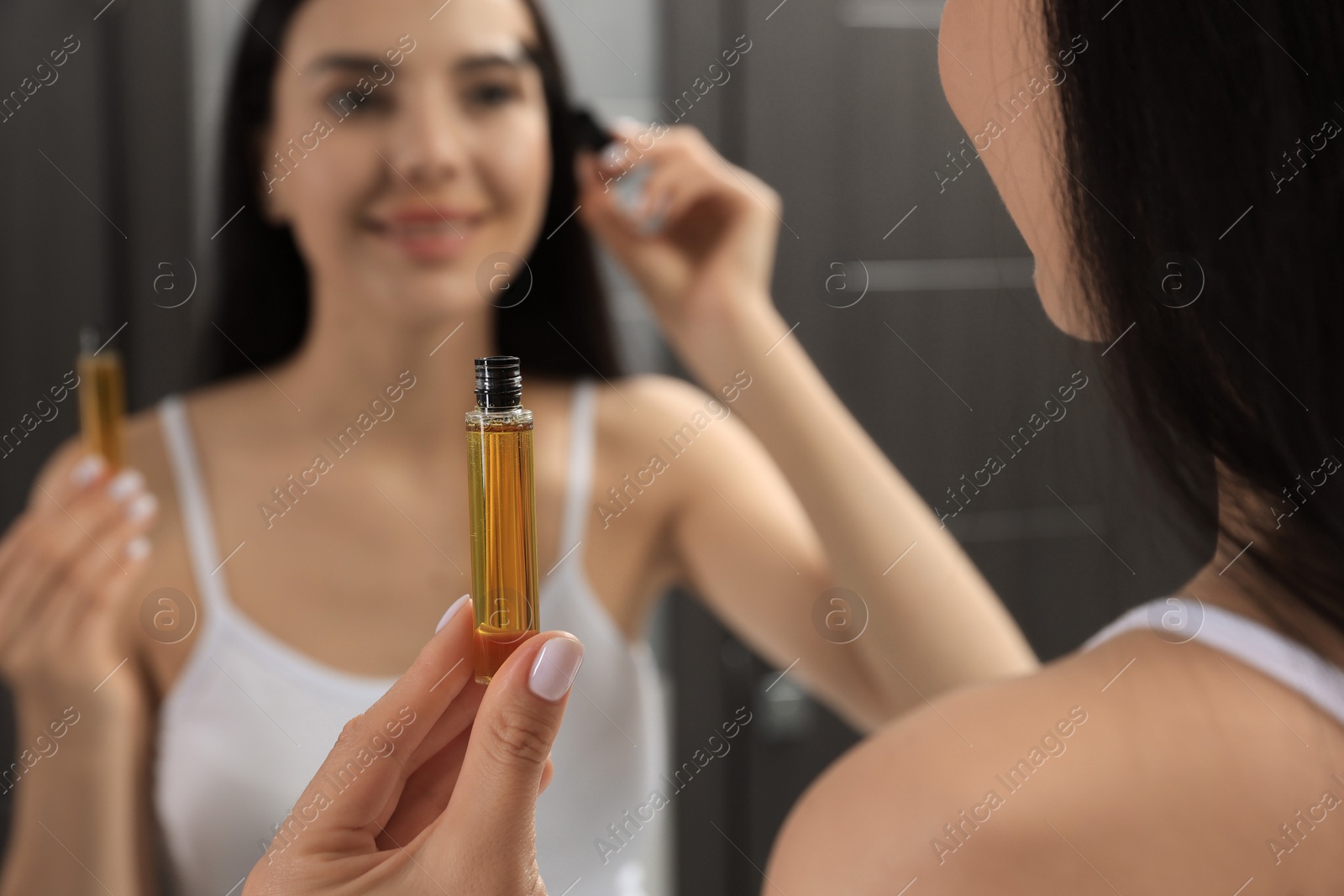 Photo of Young woman applying oil onto eyelashes near mirror indoors, closeup