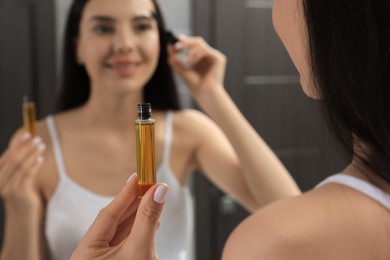 Young woman applying oil onto eyelashes near mirror indoors, closeup