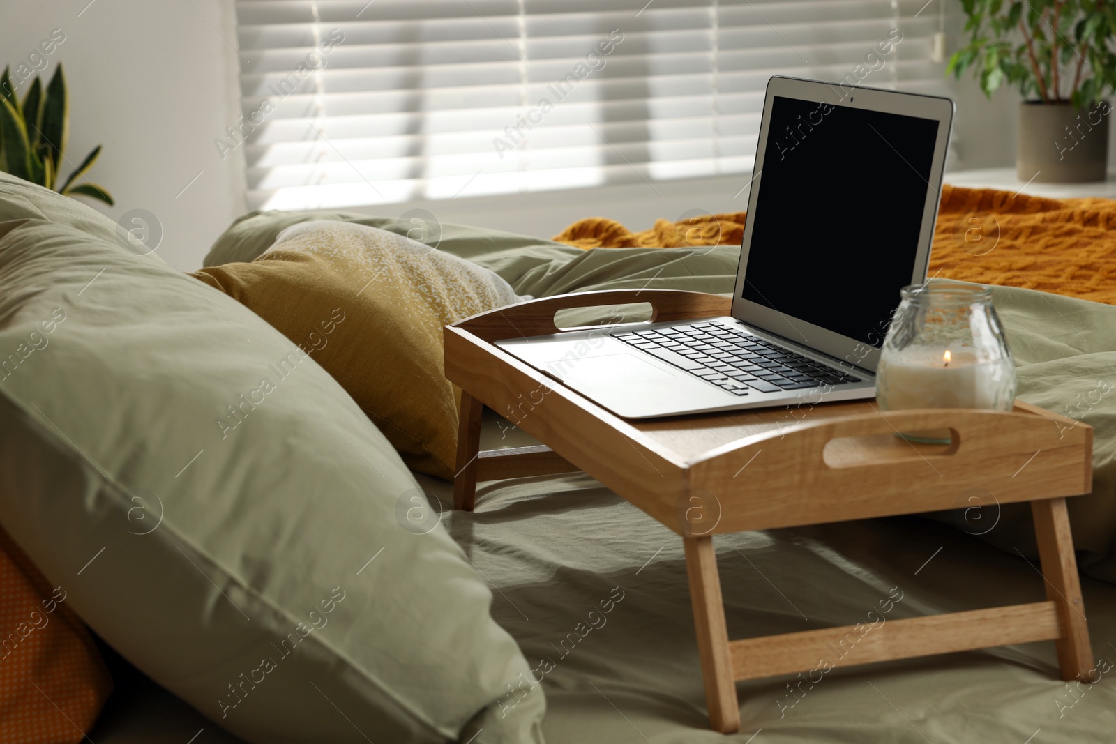 Photo of Wooden tray with modern laptop and burning candle on bed indoors