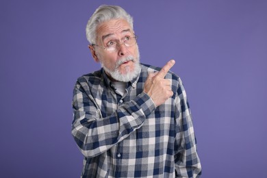 Surprised senior man pointing at something on violet background